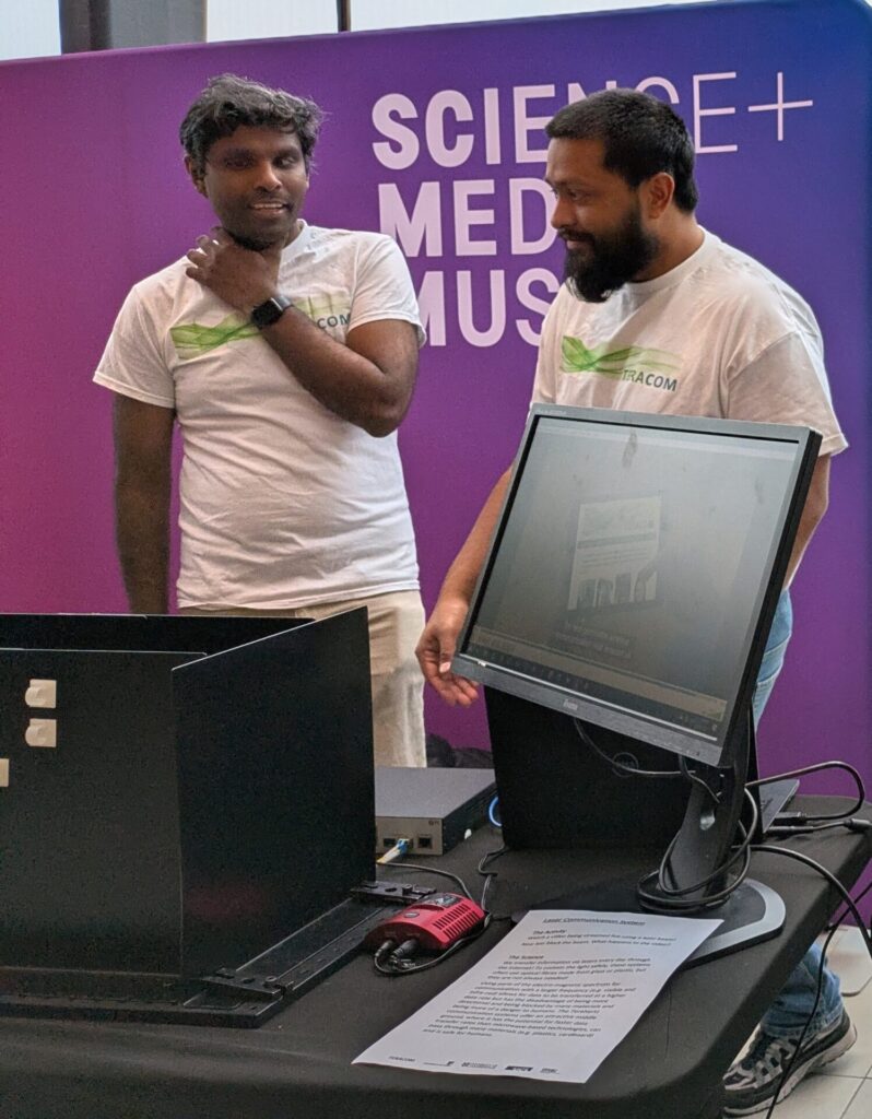 Two TeraCom researchers in 'TeraCom' t-shirts, stood in front of a sign saying 'Science + Media Museum', with a laser comms demo in the foreground.